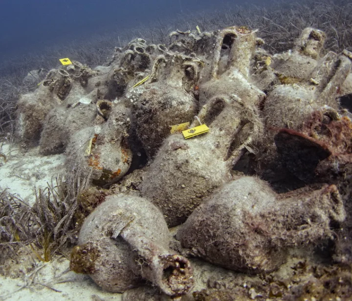 Stacked ancient amphorae from the Peristera shipwreck in Alonnisos, Greece, partially buried on the seabed with marine growth and sediment.